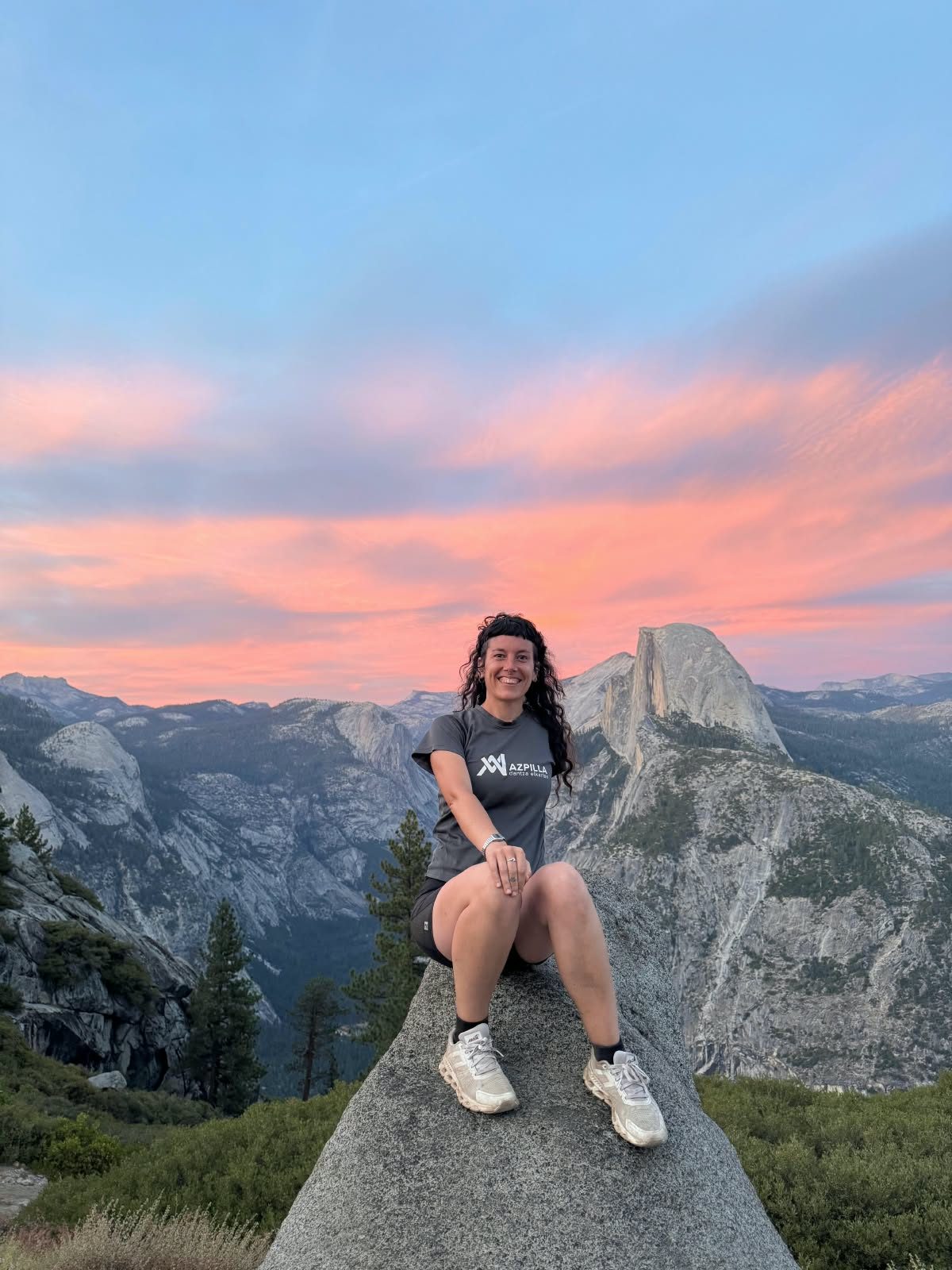 Maider irribarrez, mendian harkaitz baten gainean eserita, atzean Half Dome eta ilunabar arrosa bat duela